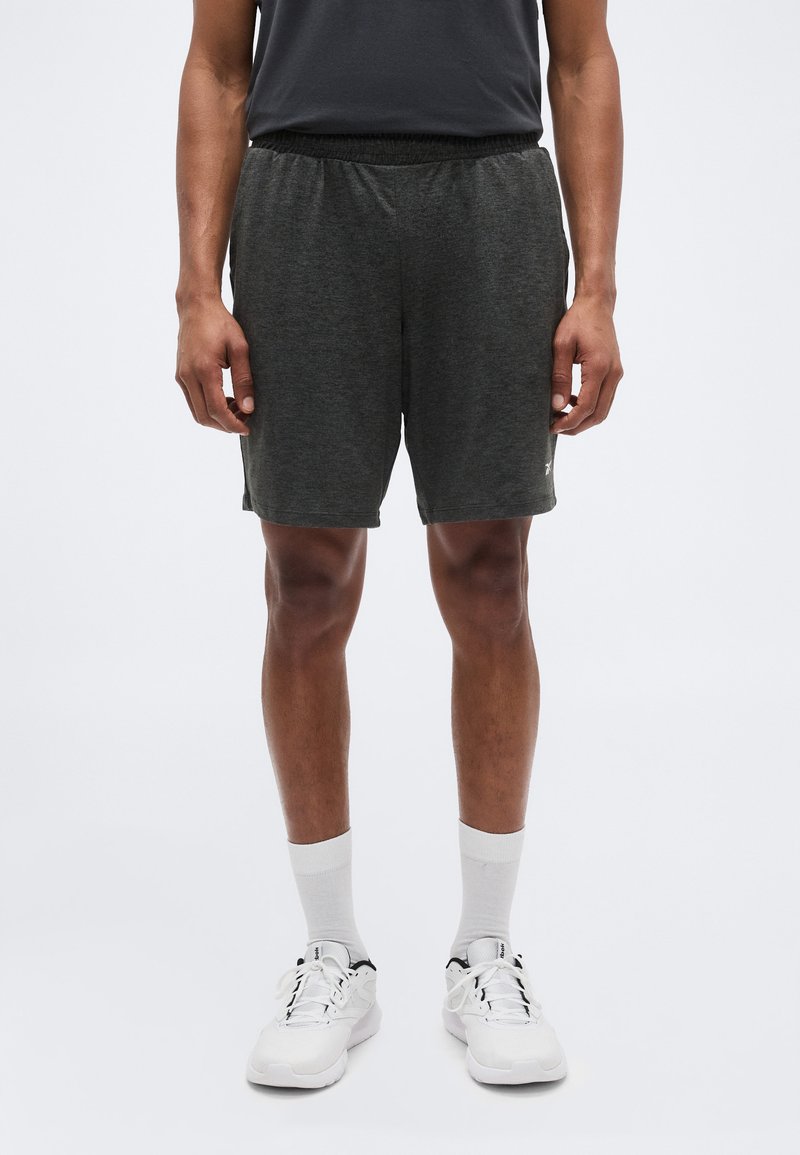 Άνδρας που στέκεται φορώντας σκούρα γκρι αθλητικά σορτς, λευκές κάλτσες μέχρι τη μέση της γάμπας και λευκά αθλητικά παπούτσια, μπροστά σε απλό φωτεινό φόντο.