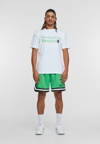 Hvid t-shirt med grøn grafisk tekst, der står "New York City Basketball," sammen med grønne mesh shorts med sorte detaljer og logo.