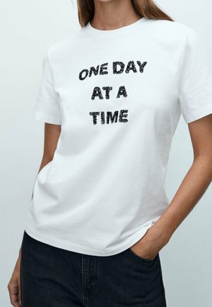 Kvinde iført en hvid t-shirt med sort teksturtekst "ONE DAY AT A TIME", stående med den ene hånd i lommen på mørke bukser.