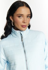 Jachetă de puf de culoare albastru deschis, cu guler înalt, textură matlasată, fermoar frontal și un design elegant, asortată cu cercei rotativi din perlă.
