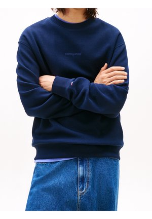 Osoba nosząca granatową teksturowaną bluzę z logo „tommy jeans” oraz niebieskie dżinsy, z założonymi rękami na białym tle.