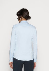 Mujer con el cabello oscuro en un moño bajo, vestida con una camisa de manga larga azul clara y pantalones oscuros, vista desde atrás sobre un fondo sencillo.
