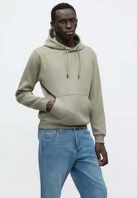 Olivengrønn hettegenser med foranlomme, snøring i hetten og ribbestrikkede mansjetter, kombinert med lyseblå denimjeans. Glatt stofftekstur.