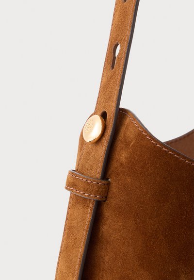 Detalles de un bolso de ante marrón que presenta una superficie texturizada, una correa plana con un botón a presión en tono dorado y costuras a lo largo de los bordes.