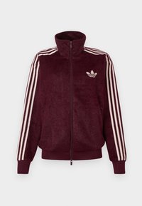 Jaqueta de desporto em bordeaux, em tecido peluche, com fecho éclair e três riscas brancas nas mangas, além do logo trefoil branco da Adidas no lado esquerdo do peito.