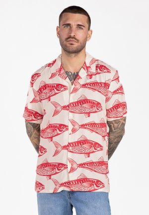 Mężczyzna z krótkimi włosami i tatuażami, ubrany w beżową koszulę z krótkim rękawem z czerwonymi wzorami ryb oraz niebieskie dżinsy na białym tle.