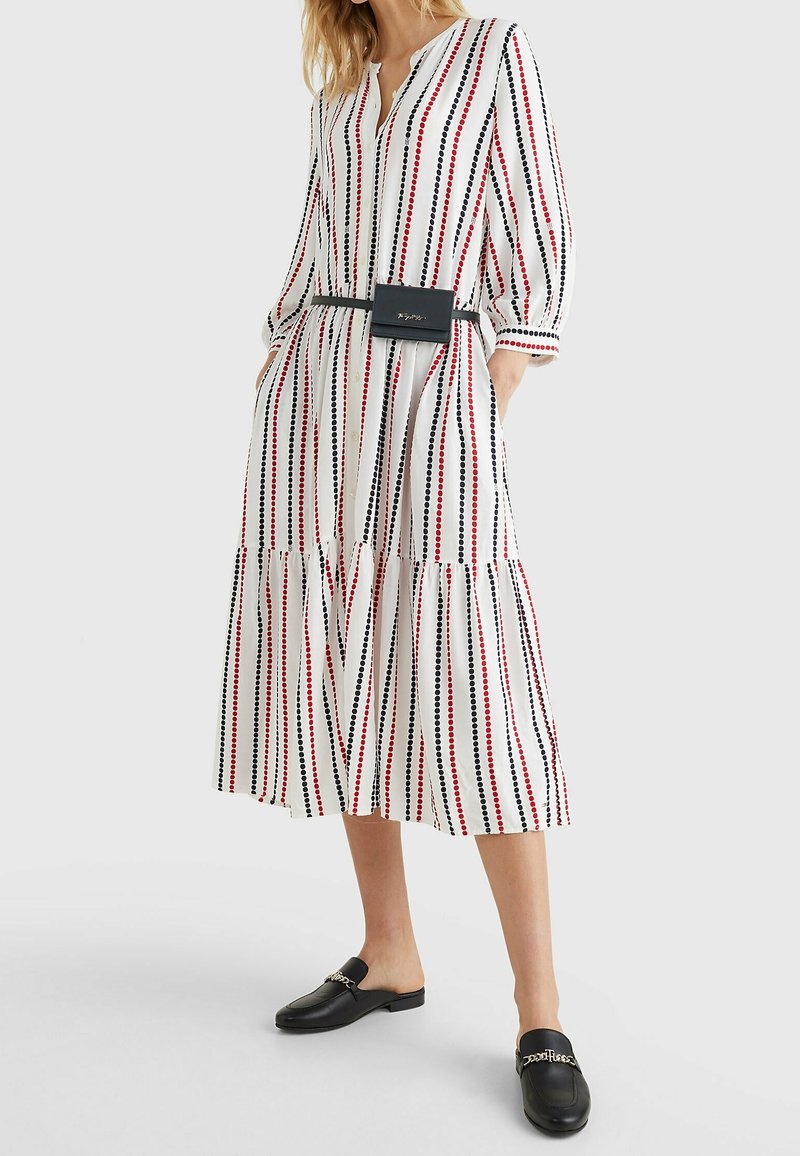 Femme portant une robe midi blanche avec des rayures verticales rouges et noires à pois, un sac ceinture noir et des mocassins noirs sans lacets avec détail chaîne.