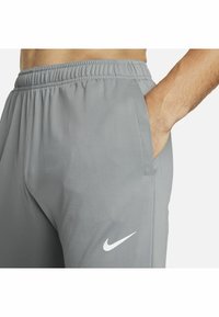 Mann in grauen Nike-Sweatpants mit elastischem Bund, Hand in der Tasche, seitliche Ansicht und Nike-Logo am Oberschenkel.