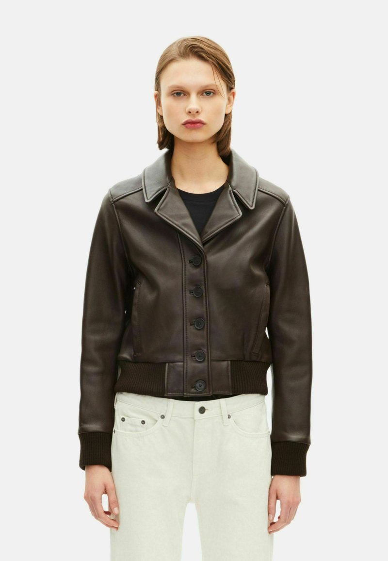 Bruine leren korte jas met een reverskraag, knopen aan de voorkant en geribde manchetten. Draagt over een zwart topje en lichte broek.