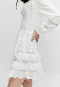 Robe blanche à volants avec trois couches, ornée de subtils accents de dentelle argentée, manches longues, et une ceinture nouée pour un ajustement modulable.