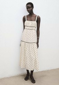 Vestido crema con patrón de puntos negros, escote cuadrado, tirantes anchos, cuerpo fruncido, falda de volantes y detalles de ribete negro.