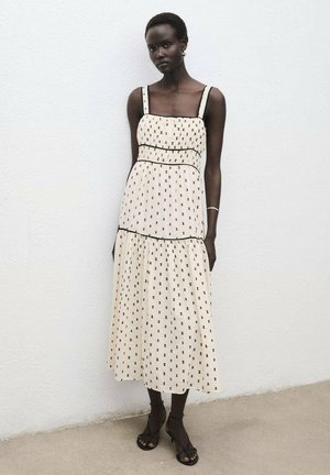 Vestido crema con patrón de puntos negros, escote cuadrado, tirantes anchos, cuerpo fruncido, falda de volantes y detalles de ribete negro.