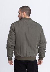 Hombre con cabello corto y barba, vistiendo una chaqueta bomber verde oliva, visto desde atrás contra un fondo claro y liso.