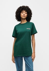 Πράσινο βαμβακερό μπλουζάκι με κοντά μανίκια, στρογγυλή λαιμόκοψη και ροζ λογότυπο. Συνδυασμένο με φαρδιά μπλε τζιν παντελόνια.