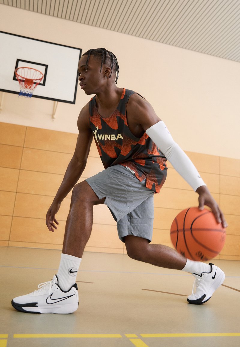 Koszykarz w czarno-pomarańczowej, wzorzystej koszulce bez rękawów i szarych szortach, drybluje z piłką do koszykówki, mając na sobie białe buty Nike.