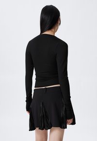 Woman with long black hair wearing a fitted black long-sleeve top and a black skirt with pleated fabric panels, standing against a plain background.