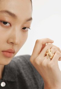 Anillo de color dorado con un diseño texturizado que recuerda a un caimán, llevado en la mano, contra un fondo neutro con luz suave.