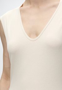 Gros plan sur une personne portant un haut sans manches beige clair avec un décolleté en V profond sur un fond uni.