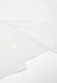 Camiseta de algodón blanca con dobladillo recto, bordes ligeramente deshilachados y detalles de Velcro para un ajuste seguro en las esquinas inferiores.