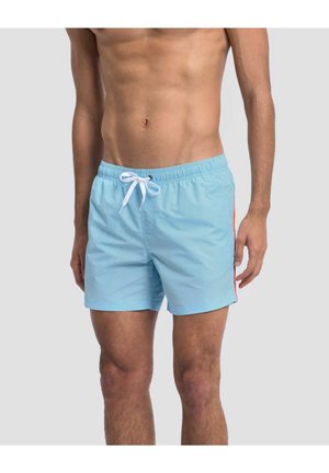Mann trägt hellblaue Badeshorts mit weißem Kordelzug und orangen Seitenstreifen, steht vor einem schlichten hellen Hintergrund.