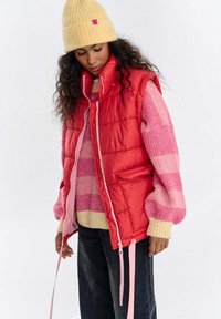 Czerwona kamizelka puchowa z różowymi akcentami i bocznymi kieszeniami, noszona na różowo-kremowym swetrze w paski. Do tego żółta dzianinowa czapka.