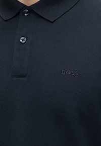 Mørk marineblå polotrøye med to knapper kneppet, med en liten brodert "BOSS"-logo på venstre brystside.