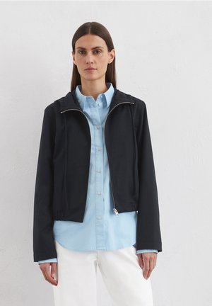 Black cropped jacket with a zip front, featuring a hood and long sleeves. Worn over a light blue button-up shirt. Smooth texture.
