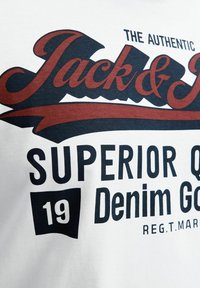 Hvid bomulds t-shirt med bold sort og rød grafisk tekst, der siger "Jack & Jones," "Superior Quality," og "Denim Goods," med et lille tal "19."