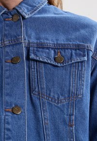 Giacca di jeans blu con taglio classico, dotata di due tasche a toppa, bottoni in bronzo e dettagli di cucitura a contrasto. Tessuto testurizzato visibile.