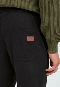Black cotton pants featuring a back pocket with a brown leather logo patch. The waistband is elastic, and the texture is soft and smooth.