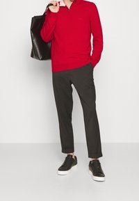 Pull rouge à fermeture éclair avec col blanc, pantalon foncé et baskets noires. Tenant un grand sac fourre-tout noir. Textures de tissu lisses.