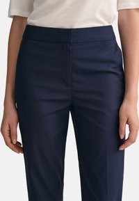 Tamsiai mėlyni kelnes iš glotnaus audinio, vidutinio aukščio liemuo, pritaikytas siluetas, šoninės kišenės ir švarus priekio dizainas.