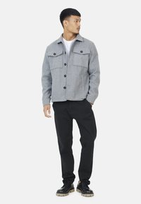 Grijze wollen jas met twee borstzakken, knoopsluiting en een opgestikte kraag, gecombineerd met zwarte broek en zwarte schoenen. Getextureerde stof.