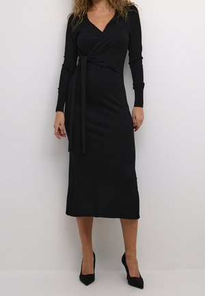 Woman wearing a long black wrap dress with long sleeves and black high heels, standing against a plain white background.