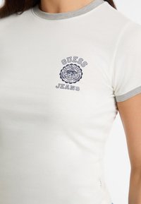 Balta prigludusi marškinėliai su pilku apykakle, priekyje yra mėlynai siuvinėtas logotipas, kuris sako "GUESS Jeans Established 1981".