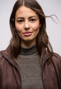 Giacca marrone con cerniera sopra un dolcevita grigio scuro a coste. Presenta texture morbide e una vista ravvicinata, con capelli che scorrono delicatamente.