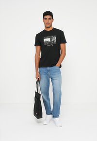 Camiseta de algodón negra con un estampado gráfico que dice "TOMMY JEANS" y "Nueva York". Vaqueros de mezclilla azul claro y zapatillas blancas. Bolsa tote negra.