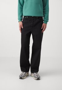 Svarta jeans i denim med avslappnad passform, fem fickor och metalldetaljer, kombinerade med en grön hoodie och gråa sneakers.