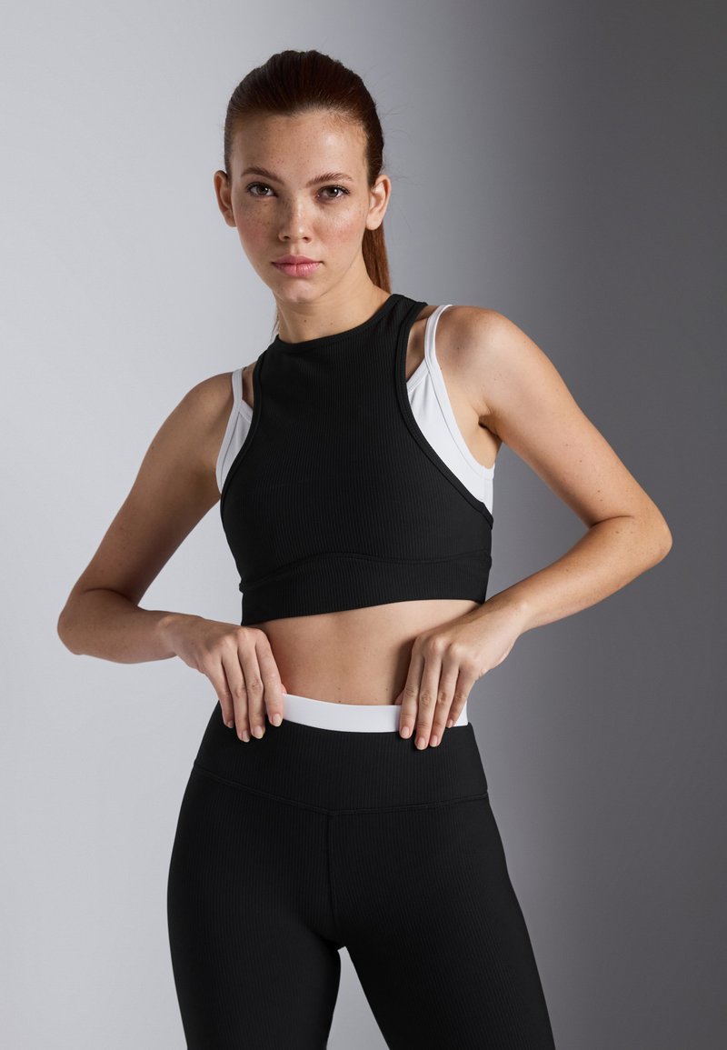 Musta ribbed crop-top korkean kaula-aukko kanssa valkoisen urheiluliivin päällä. Mustat korkea vyötäröiset legginsit, joissa on valkoinen vyötärö.