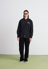 Camicia nera con bottoni a maniche lunghe, dotata di due tasche sul petto e logo ricamato, abbinata a pantaloni neri e occhiali da sole. Texture liscia, design casual.