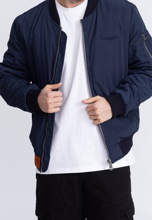 Homme portant un blouson aviateur bleu marine sur un t-shirt blanc et un pantalon noir, tenant la veste ouverte près de la poitrine, haut du corps visible.