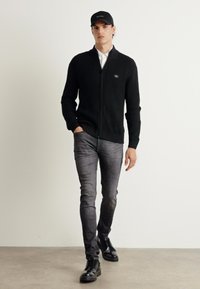 Maglione zip-up a coste nero, camicia bianca con colletto, jeans grigi slim-fit e stivali neri stringati, abbinati a un berretto nero.