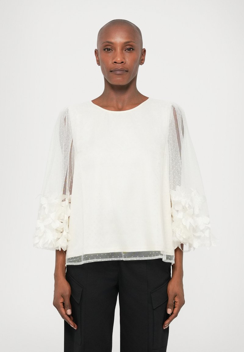 Bela majica s kratkimi, prosojnimi pufastimi rokavi, okrašenimi s cvetličnimi detajli na zapestjih. Gladka tkanina z pikčasto teksturo.