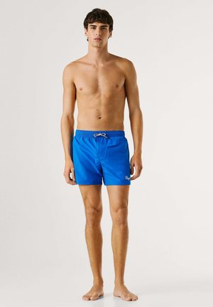 Mladić stoji bosi, nosi svijetloplave kupaće gaće s bijelim vezicama, na jednobojnoj pozadini.