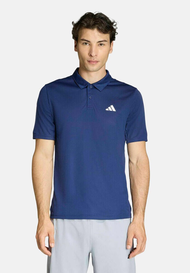 Jeune homme debout, portant un polo Adidas bleu marine et un pantalon gris clair, sur un fond uni.