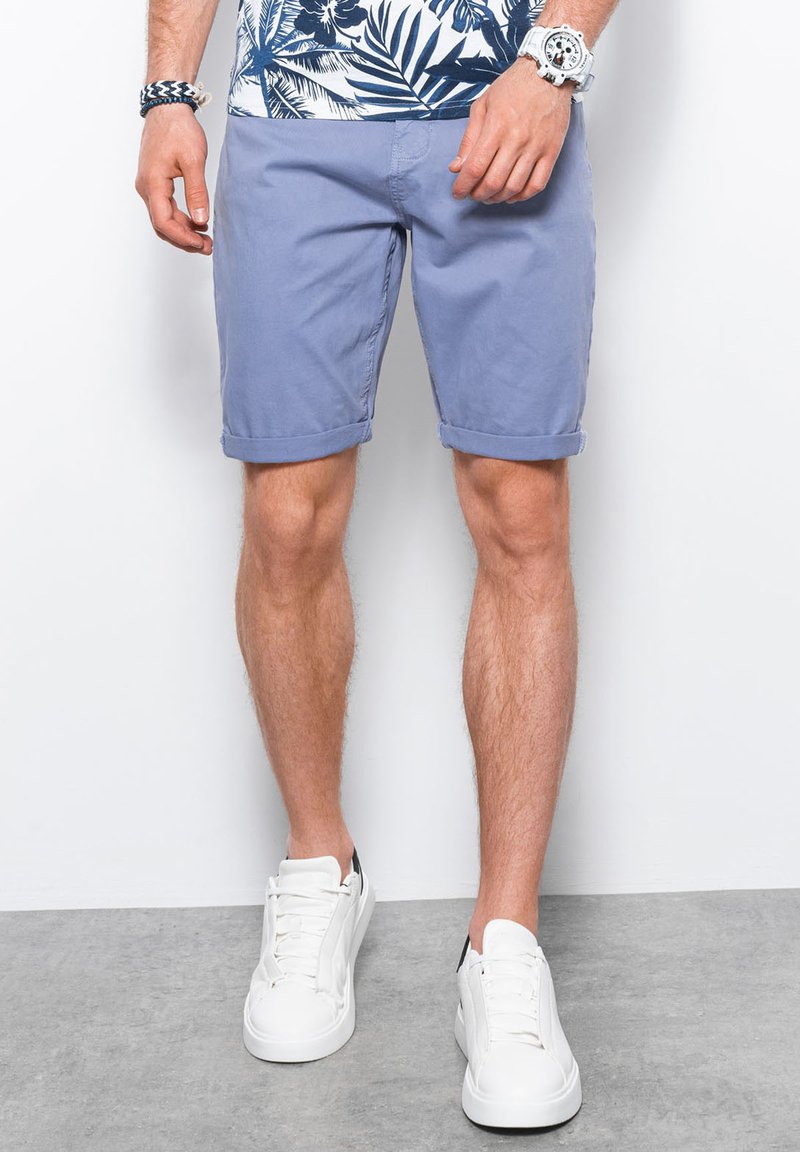 Pantaloni corti in cotone blu chiaro con orli arrotolati, abbinati a sneakers bianche. I pantaloni corti hanno una vestibilità rilassata e una texture liscia.
