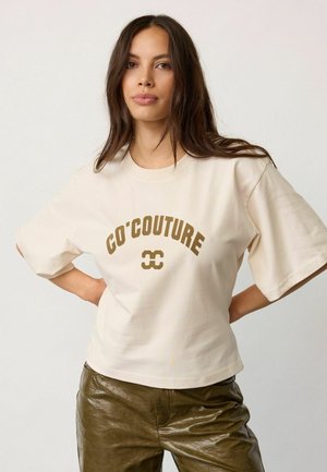 Junge Frau mit langen dunklen Haaren trägt ein beiges T-Shirt mit dem Text "CO'COUTURE" und olivgrünen Lederhosen, posiert mit den Händen in der Hüfte.