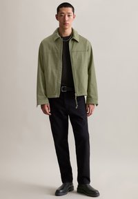 Grüne Baumwolljacke mit Reißverschluss, Fronttaschen und weitem Kragen, getragen über einem schwarzen Hemd und kombiniert mit schwarzen Hosen und Stiefeln.