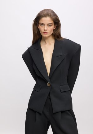Black blazer with exaggerated shoulder design, deep V-neck, single gold button, structured fit, and two front pockets. Tailored trousers complement.