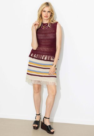 Femme portant un haut en crochet bordeaux sans manches et une jupe à franges rayée, accessoirisée avec des bijoux dorés et des sandales à plateforme noires.